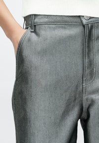 Mâna unei persoane odihnindu-se în buzunarul frontal al unei perechi de blugi gri estompați, asortați cu un top alb, cu dungi.