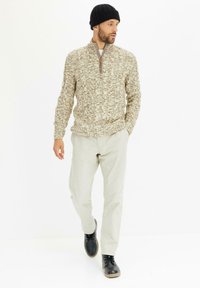 Maglione a maglia beige con texture e colletto a mezza zip, abbinato a pantaloni chiari e stivali neri con lacci, indossato da un uomo in piedi.