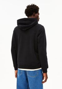 Felpa nera con cappuccio, dal taglio rilassato. Presenta polsini e orlo a coste. Indossata sopra una maglietta chiara e abbinata a jeans blu.