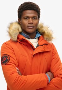 Chaqueta parka naranja brillante con capucha de piel sintética, que presenta un parche de logo en la manga y bolsillos con cremallera. Capa sobre un suéter crema.