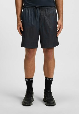Mann trägt schwarze Sportshorts, schwarze Socken mit "BOSS"-Aufschrift und schwarze Turnschuhe, steht vor einem schlichten hellen Hintergrund.