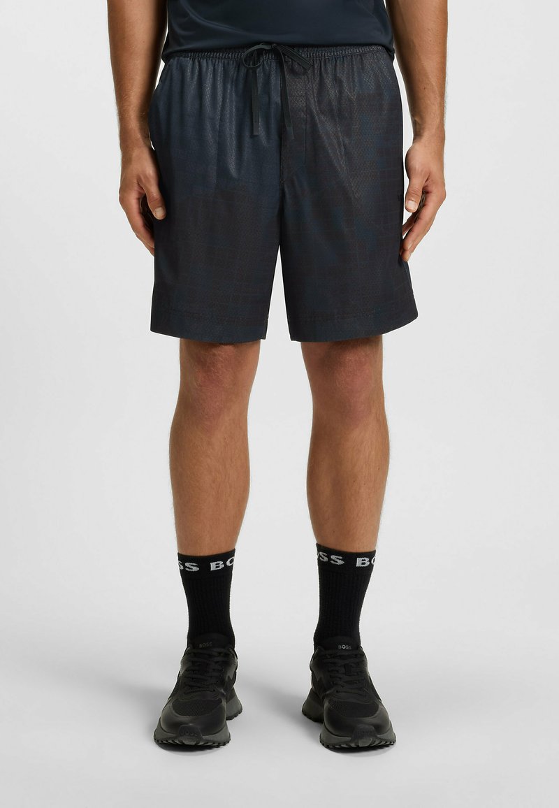 Mężczyzna w czarnych szortach sportowych, czarnych skarpetach z napisem "BOSS" oraz czarnych sneakersach stoi na tle jednolitego jasnego tła.