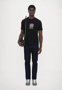 Camiseta de algodón negra con un estampado gráfico colorido, jeans azul oscuro y zapatillas color teal. El modelo lleva un pequeño bolso negro cruzado.