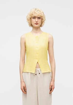 Person med kort krøllete blondt hår iført en gul ermeløs knappet topp og lyse beige bukser med snor, stående foran en ensfarget bakgrunn.