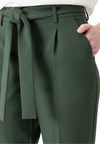 Un pantalon tailleur vert présente une ceinture en tissu, des poches latérales, des plis et une texture lisse, mettant en avant un design structuré.