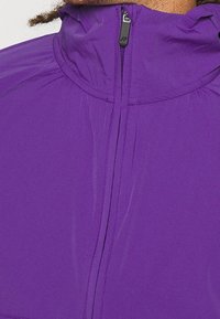 Giacca leggera viola con colletto alto, dotata di zip frontale e dettagli arricciati vicino al collo. Texture liscia.