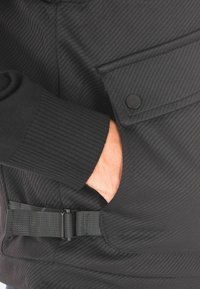 Giacca nera con zip realizzata in un tessuto a coste testurizzato, con un taschino con bottone e un dettaglio con cinturino regolabile di lato.