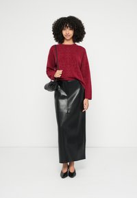 Μπορντό πουλόβερ με υφή, μαύρη φούστα maxi από δέρμα, μαύρη τσάντα και παπούτσια με μυτερή μύτη, στέκεται μπροστά σε απλό λευκό φόντο.