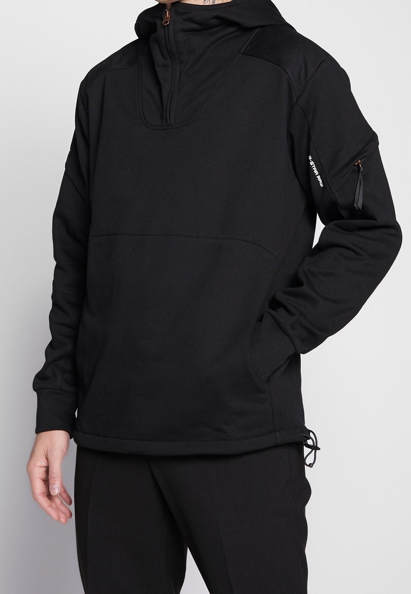 Černý kapucňový pulover vyrobený z jemné tkaniny; má poloviční zip vpředu, boční kapsy a texturované ramenní panely s tažným lemem.