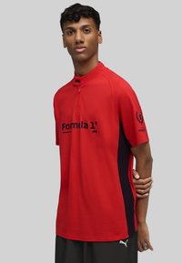 Camisa polo roja con cuello alto, con acentos laterales negros y el logo de "Formula 1" en la parte delantera. Fabricada en tejido suave, de mangas cortas.