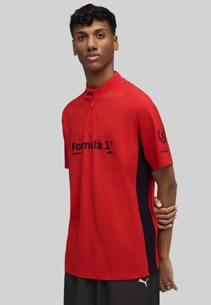 Rotes Polohemd mit hohem Kragen, ausgestattet mit schwarzen Seitenelementen und dem "Formula 1"-Logo auf der Vorderseite. Aus glattem Stoff, mit kurzen Ärmeln.