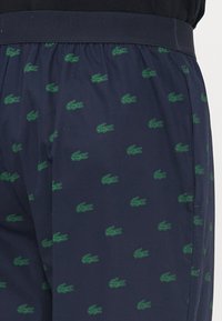 Marineblå shorts med grønn krokodillelogo-mønster. Elastisk midje og glatt tekstur. Har vekslende små logoer gjennom hele stoffet.