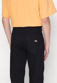 Pantalones de mezclilla negros con corte recto, que cuentan con dos bolsillos traseros y un pequeño parche con el logo de la marca. Combinados con una camiseta de color naranja claro.