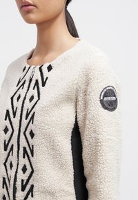 Kremowa kurtka polarowa z czarnymi geometrycznymi wzorami, zapięciem na zamek błyskawiczny i teksturowaną powierzchnią. Czarne akcenty materiałowe po bokach oraz naszywka z logo.