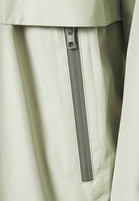 Ljusgrön jacktyg med en slät struktur, som har en kontrasterande grå dragkedjedetalj. Dragkedjan är centralt placerad och funktionell.