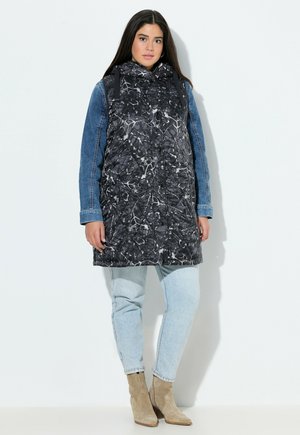 Kvinde stående iført en sort mønstret ærmeløs hættetrøje over en blå denimjakke, lysevaskede jeans og beige ruskindsankelstøvler.