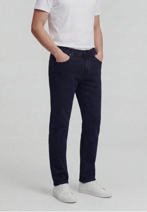 Person wearing dark navy slim-fit jeans, white t-shirt, and white sneakers, standing with hands in pockets against plain background.