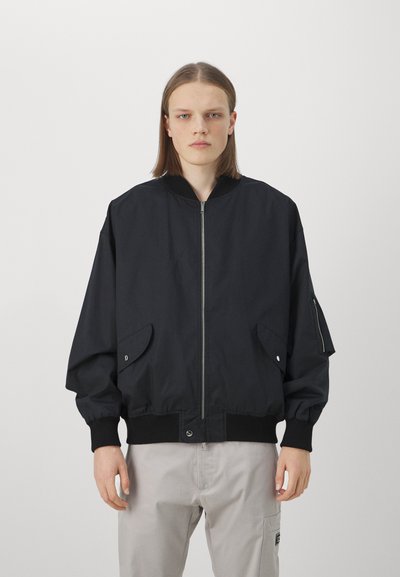 Giacca bomber nera con chiusura a zip, polsini a costine e due tasche frontali. Presenta un collo arrotondato e una vestibilità comoda.