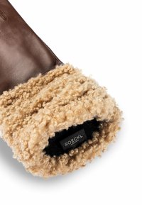 Guante de piel marrón con un puño de shearling de color canela y esponjoso. Presenta una textura suave y una etiqueta de marca visible en el interior.