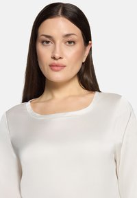 Hvid bluse med en glat, satinlignende tekstur, der har en rundet halsudskæring og lange ærmer. Stoffet falder blødt.