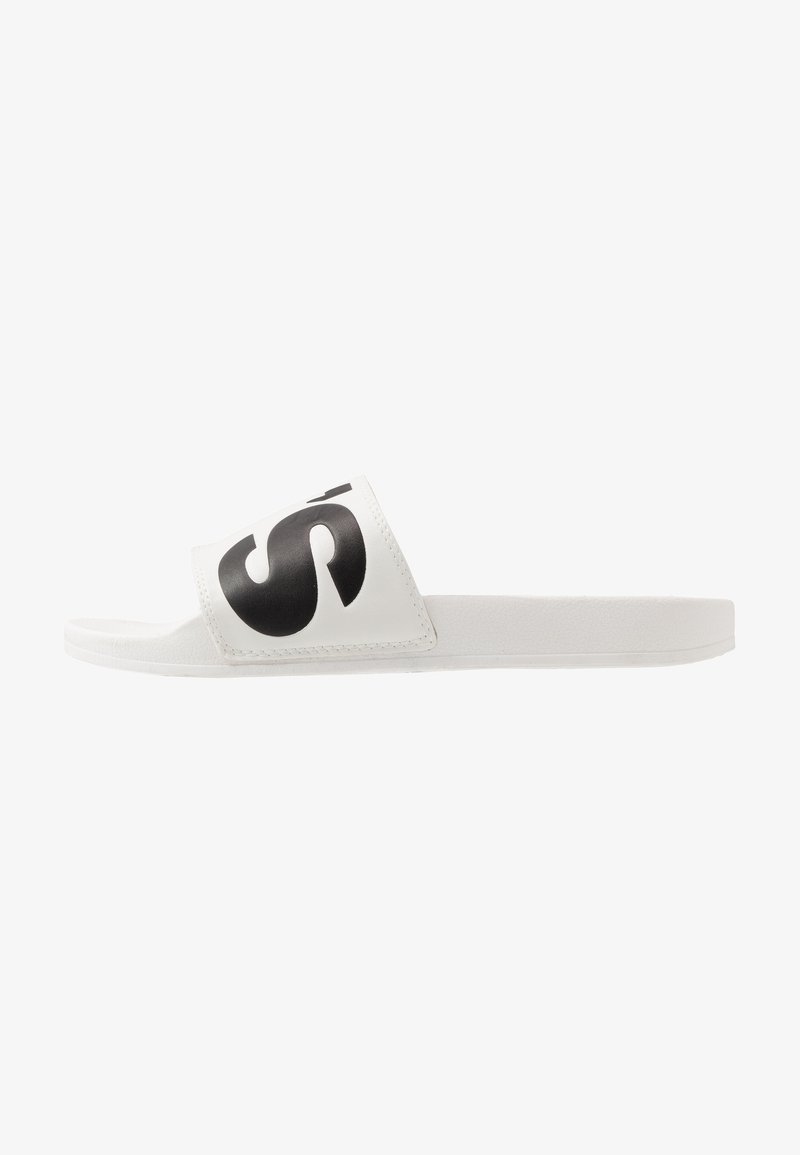 Sandale blanche avec une large sangle arborant un grand logo noir. Matériau doux, semelle plate, design minimaliste et texture lisse.