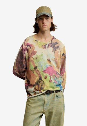 Noormees, kes kannab mitmevärvilise lillemustriga dressipluusi, helerohelisi pükse, pruuni vööd ja khaki värvi pesapalli nokamütsi, seisab valge tausta ees.