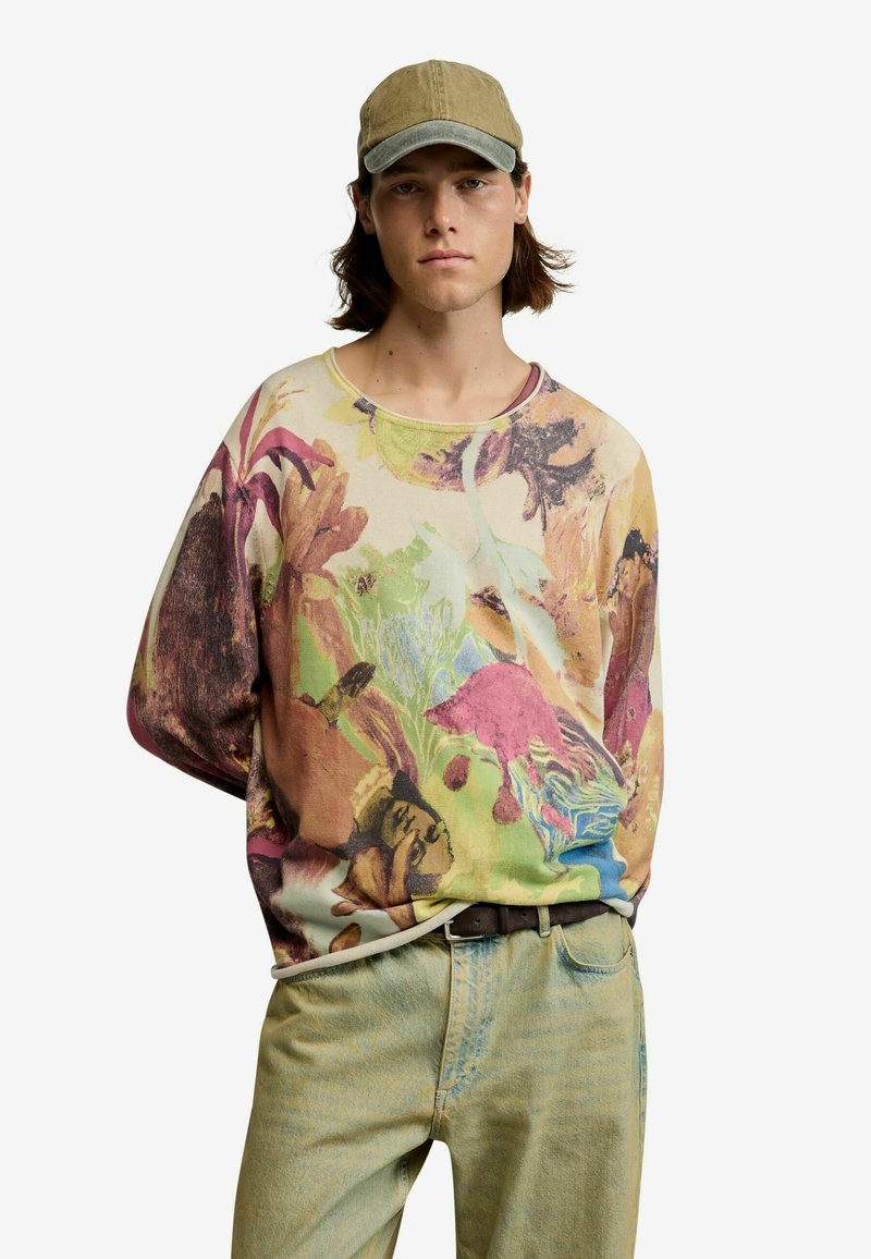 Young man wearing a multicolored floral-print sweatshirt, light green pants, a brown belt, and a khaki baseball cap, standing against a white background.