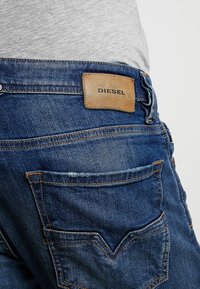 Bakomvy av blå denimjeans med sömnadsdetaljer och en brun läderlapp märkt "DIESEL" ovanför den högra bakfickan.