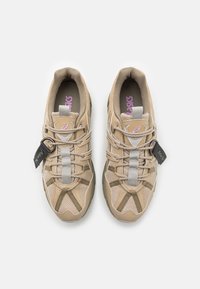 Béžové sportovní boty vyrobené z prodyšných a syntetických materiálů, s šedými akcenty, elastickým uzávěrem a štítkem připevněným na tkaničkách.