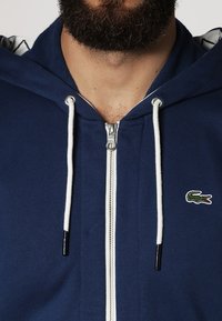 Mörkblå zip-up hoodie i bomullsblandning, med en vit dragsko, silverfärgad dragkedja och liten grön krokodil logotyp på bröstet.