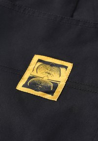 Tissu noir avec une étiquette rectangulaire jaune affichant un motif graphique noir de deux formes courbes. Étiquette cousue dans un coin.