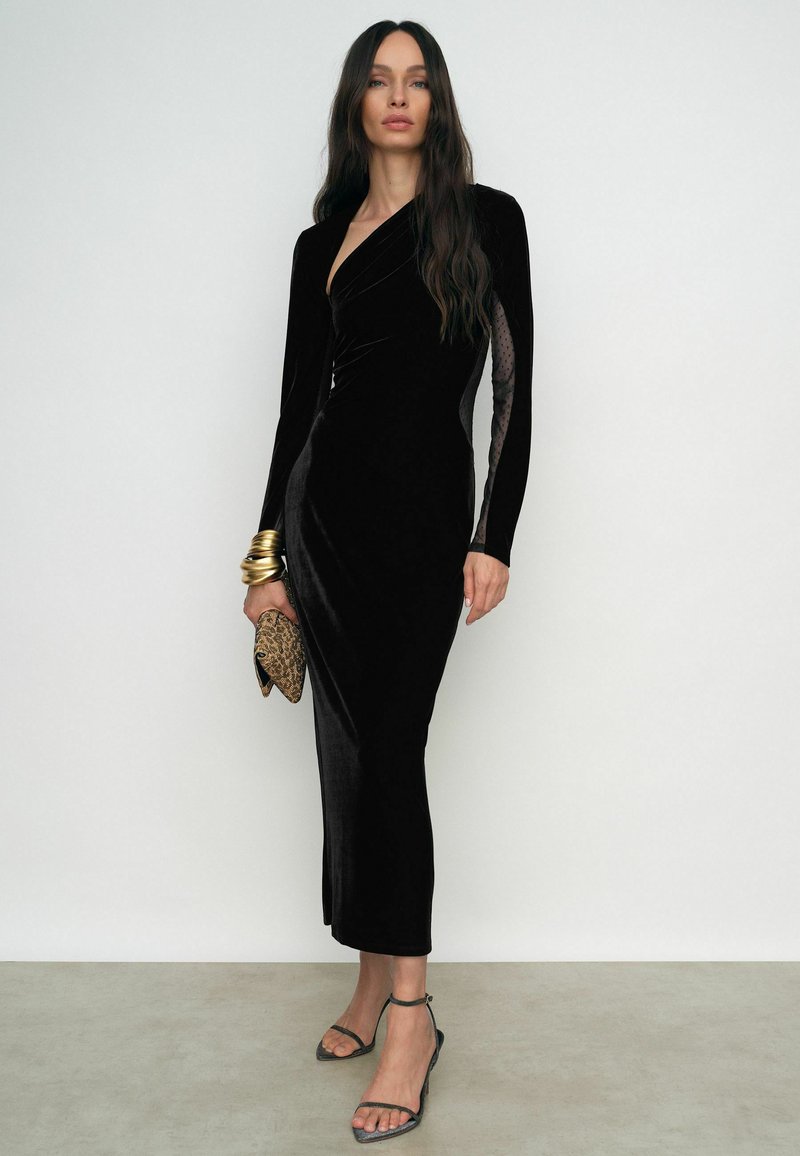 Melnas samta kleitas ar garām piedurknēm, apvērsta dizaina, izšūtām caurspīdīgām sānu daļām un augstas šķēles. Valkāta ar zelta aprocēm un rakstainu somiņu.