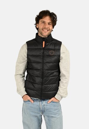 Homme souriant portant un sweat-shirt gris clair, un gilet sans manches matelassé noir et un jean bleu clair, les mains dans les poches.