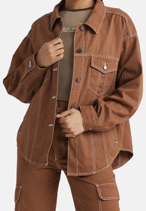 Chaqueta de algodón marrón con costuras blancas, con cierre de botones, dos bolsillos en el pecho y un corte relajado, combinada con una blusa ajustada.