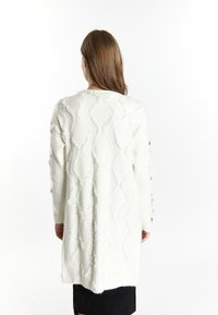 Pullover bianco lungo con un motivo a diamante texture, bordi sfrangiati e dettagli con bottoni sulle maniche. Materiale in maglia leggero.
