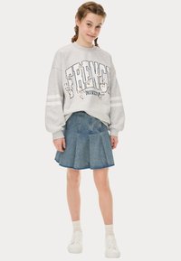 Sweatshirt gris avec l'imprimé "FRENCH RIVIERA", associé à une jupe plissée en denim. Des baskets blanches et des chaussettes de couleur claire complètent le look.