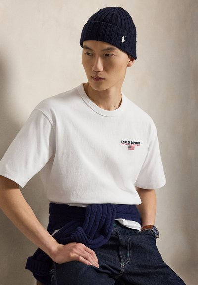 Egy férfi, aki sötétkék kötött sapkát, fehér Polo Sport pólót, sötétkék pulóvert kötve a derékra és sötét farmert visel, egyszerű háttér előtt ülve.