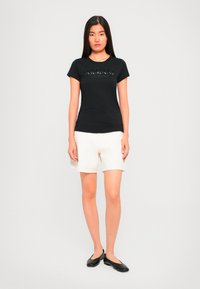 Ung kvinne står, iført svart Armani Exchange T-skjorte, hvite shorts og svarte leiligheter mot en ensfarget lys bakgrunn.