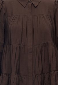 Robe marron à étages avec manches longues, fermeture boutonnée à l'avant et col pointu, montrant la texture du tissu et les détails des coutures froncées.