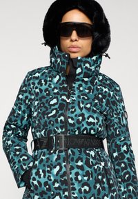 Πράσινη λεοπάρ γούνα σακακι puffer με γούνα στην κουκούλα, μαύρη ζώνη με λογότυπο και κλείσιμο με φερμουάρ. Λείο υφαντό, εφαρμοστό σχέδιο και τσέπες με φερμουάρ.