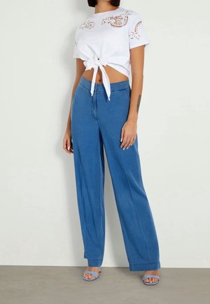 Bílý krátký top se zavazováním a motivy laserových výřezů, v kombinaci se širokými modrými džínovými kalhotami a světle modrými sandály.