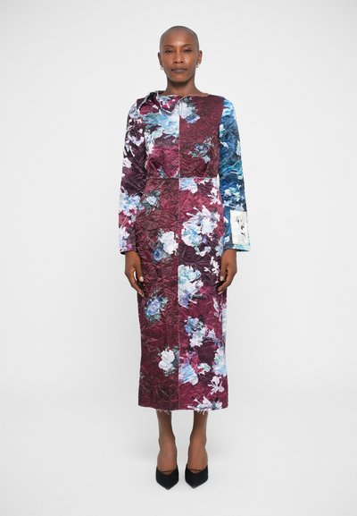 Robe longue matelassée aux motifs floraux bordeaux et bleus ; elle présente des manches longues, un décolleté rond et une silhouette droite. Des talons noirs complètent le look.