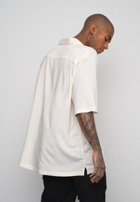 Camisa de manga corta, color crema, con una textura sutil, un collar y aberturas laterales. Presenta un corte suelto y se lleva al revés.
