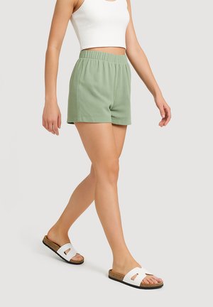 Pantaloni corti verde chiaro, con texture e vita elastica, dotati di tasche laterali. Indossati con sandali bianchi a scivolo e un crop top aderente bianco.