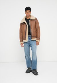 Giubbotto in pelliccia shearling marrone con colletto e polsini color crema, dotato di una cerniera frontale e cuciture scure a contrasto, abbinato a jeans azzurri chiari.