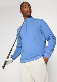 Pullover azzurro chiaro con zip a quarto, realizzato in tessuto morbido, presenta il logo Nike sulla manica. Abbinato a pantaloni beige e tiene in mano una mazza da golf.