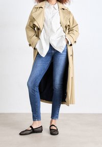 Gabardina beige, camisa blanca de rayas con botones, jeans ajustados azules, bailarinas de malla negra con tirante. Tejido suave y diseño moderno.