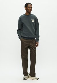 Dunkelgrauer Sweatshirt mit einem cremefarbenen Logo auf der Brust, kombiniert mit braunen geraden Hosen und neutralen Sneakern. Glatte Texturen überall.