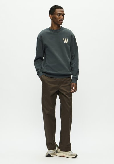 Un sweat gris foncé avec un logo crème sur la poitrine, assorti à un pantalon droit marron et des baskets de teintes neutres. Textures lisses tout au long.