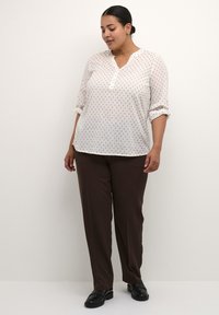 Blusa blanca con patrón de puntos azul marino, cuello con muesca y mangas enrolladas, combinada con pantalones marrones y mocasines negros. Tejido suave, corte relajado.